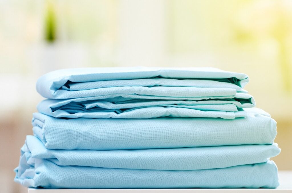 Close-up of blue clean bedding on a blurred background. A stack of folded new bed sheets on the table. Sunlight from the window.